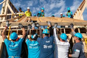 A group of volunteers work together to lift up the frame of a house while more volunteers stand on top of the roof pulling the frame up.