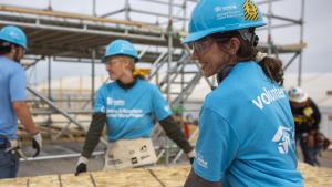 Two women in Habitat volunteer shirts carry a sheet of plywood on the 2024 Carter Work Project build site.