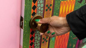 A hand with green painted nails pulls open a door with an illustrated image on it. The frame of the door is pink.