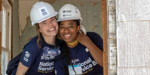 Two volunteers smile and hug on a build site. They are wearing navy blue shirts and white hard hats.