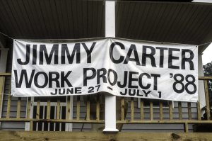A large white banner hangs on the front porch of a home. The black text says "Jimmy Carter Work Project '88 June 27-July 1"