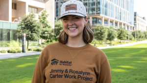 Young woman smiling wearing a tan sweatshirt and white hat, both branded with Habitat for Humanity logo and "Jimmy & Rosalynn Carter Work Project"