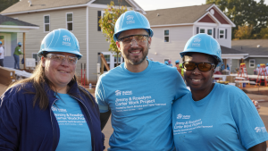 Two female volunteers and one male volunteer pose together smiling with a house construction site in the background. They are wearing light blue hardhats and matching blue T-shirts branded with the Habitat logo and "Jimmy & Rosalynn Carter Work Project"