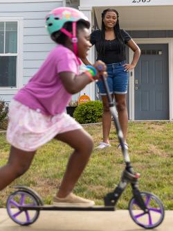 Child rides a scooter while their mom watches from the yard.
