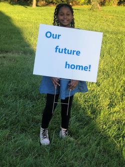young girl standing in grass holding a sign that says "our future home"