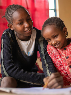 Young child sits with older sibling and watches them use a pencil on paper.