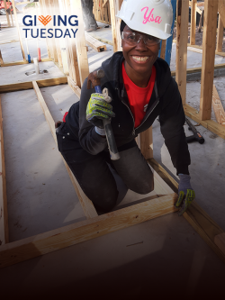 A woman wearing a white hardhat with name "Ysa" painted on it smiles at the camera while kneeling and nailing studs with a hammer on a Habitat build site. Giving Tuesday logo text on corner of image.