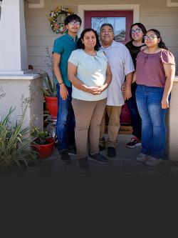 A family of five with mother, father, two older daughters and adolescent son pose smiling together on the front porch of their Habitat home.