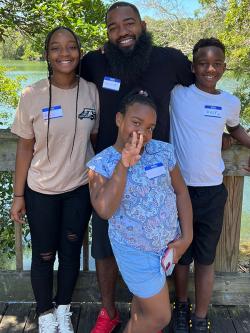 A family of four poses together standing in front of a wooden railing next to a river. The father stands in the middle of his adolescent daughter and son with his arms around them, and his younger daughter stands in front of them holding up her hand with a peace sign.