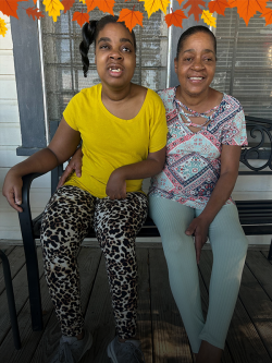 A woman smiles sitting on a porch bench with her arm around a smiling young girl who has a visible disability affecting her arm and eyes.