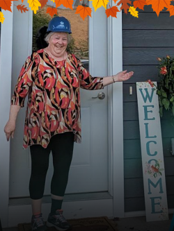 An older woman wearing a Habitat hard hat poses smiling on the front porch of her Habitat home showing off a welcome sign. A decorative graphic border of fall leaves lines the top of the image.