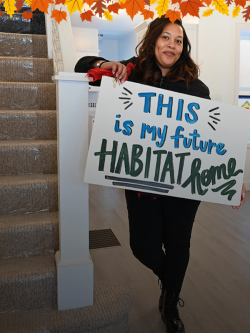 A woman poses next to a staircase inside a house with a hand-painted poster that reads "This is my future Habitat home!"