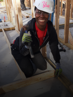 A woman wearing a white hardhat with name "Ysa" painted on it smiles at the camera while kneeling and nailing studs with a hammer on a Habitat build site.