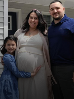 A mother and father pose in front of their home with their young daughter hugging her mother's pregnant belly.