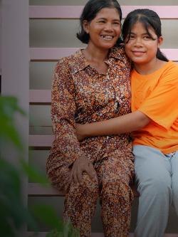 A woman with short black hair and a red floral patterned dress sits beside her daughter who is in an orange shirt and is hugging her.
