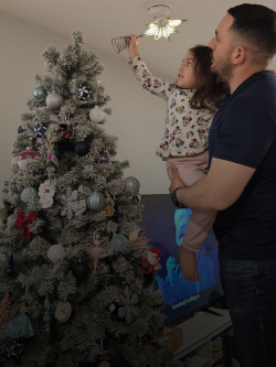 A man holds his young daughter up to place a star on top of their Christmas tree.