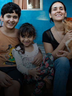 A mother holding a puppy smiles and laughs with her two children while sitting on the front steps of their bright blue home.