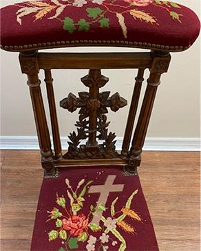 Ornate pray kneeler with red embroidered seating of a cross and flowers and the front wooden piece is carved into a cross.