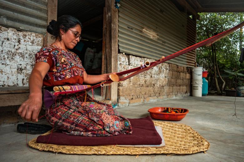 Woman in red clothing kneeling on mat while weaving 