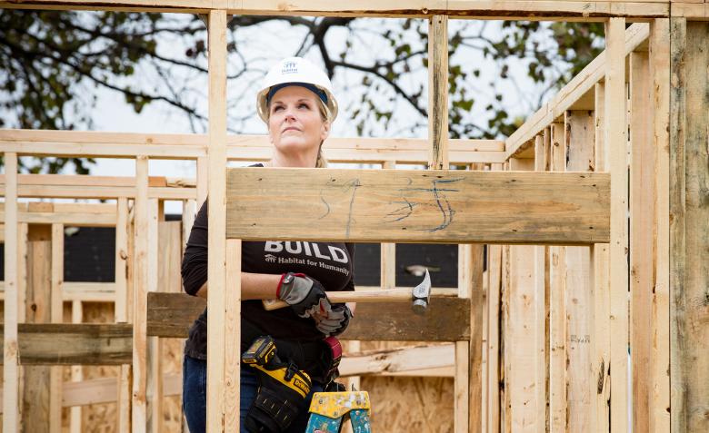 Trisha on the build site, holding a hammer and working inside the lumber walls of an unfinished house.