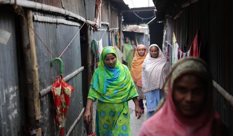 Women in colorful clothing walk down an alleyway in an informal settlement in Bangladesh.