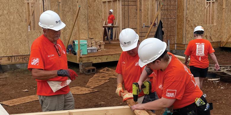 Volunteers in hard hats and orange Team Depot shirts work together on house framing at build site