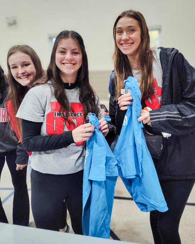 Three female student volunteers smile together while holding light blue Collegiate Challenge shirts.