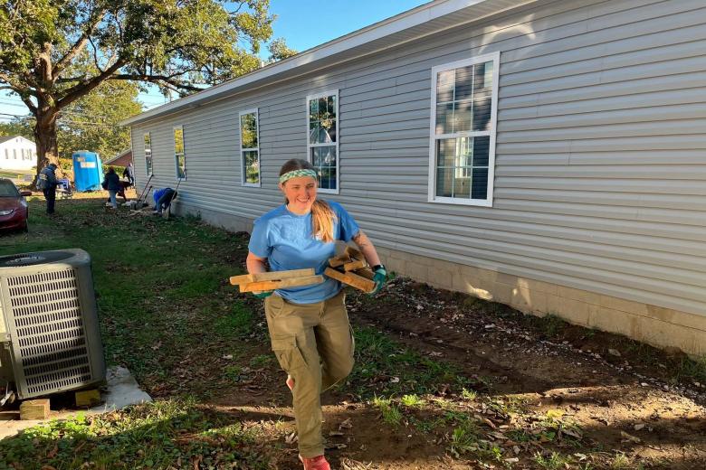 A volunteer in a blue shirt walks forward carrying pieces of lumber beside a house.