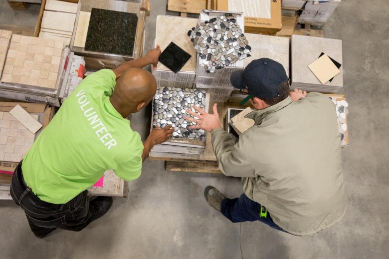 A volunteer in a neon shirt leans over a pallet of tiles to help a customer who is also leaning over the tiles and wearing a baseball cap.