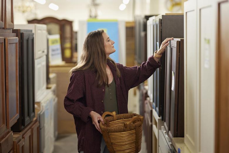 A customer stands in the middle of an aisle of cabinets in Habitat ReStore. She has one arm lifted inspecting the cabinets and the other is holding a basket.