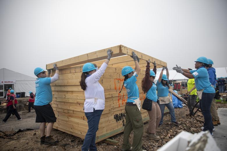 A group of Carter Work Project in bright blue Habitat hard hats work together to carry a wood wall frame.