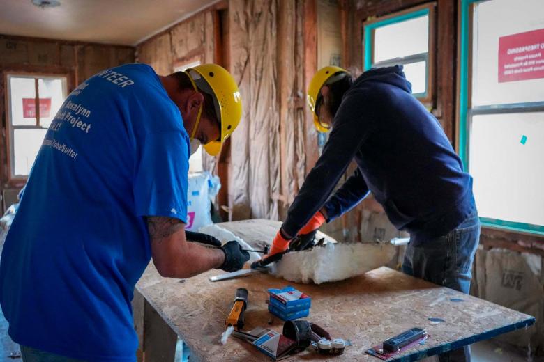Two volunteers wearing protective masks and hard hats bend over a table to work on insulation.