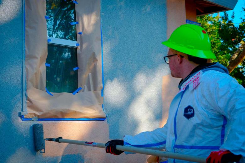 A volunteer wearing protective plastic suit and hard hat paints the exterior wall of a house with a roller brush on a pole.