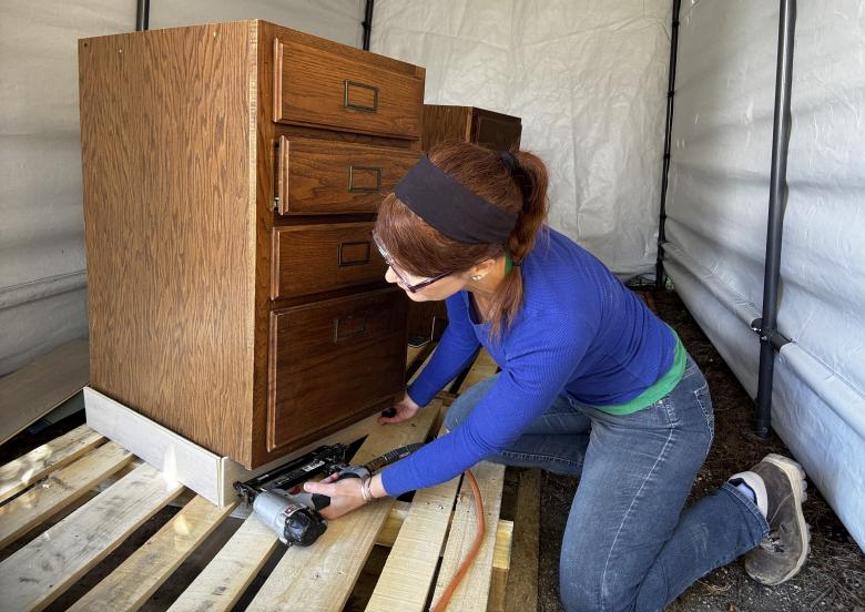 Woman in blue shirt and jeans adding trim to the bottom of a brown wood cabinet.