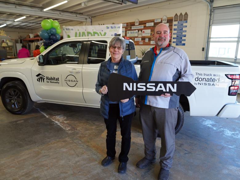 A man and woman pose together holding a key-shaped poster that says Nissan in front of a white Habitat-branded Nissan pickup truck.