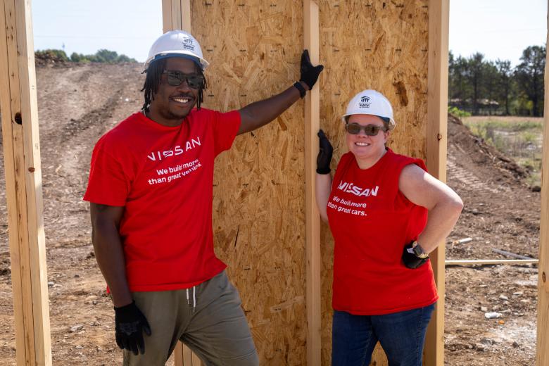 A man and woman pose smiling together on a construction site wearing matching white hardhats and red Nissan volunteer shirts.