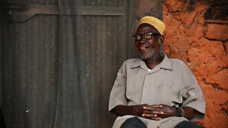 elderly man sat outside a mud home, behind him a door with netting