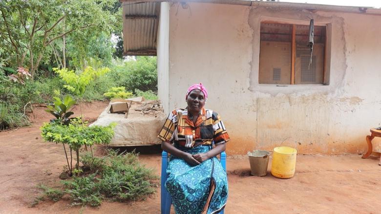 Woman sits outside a mud home, the home has a window with netting